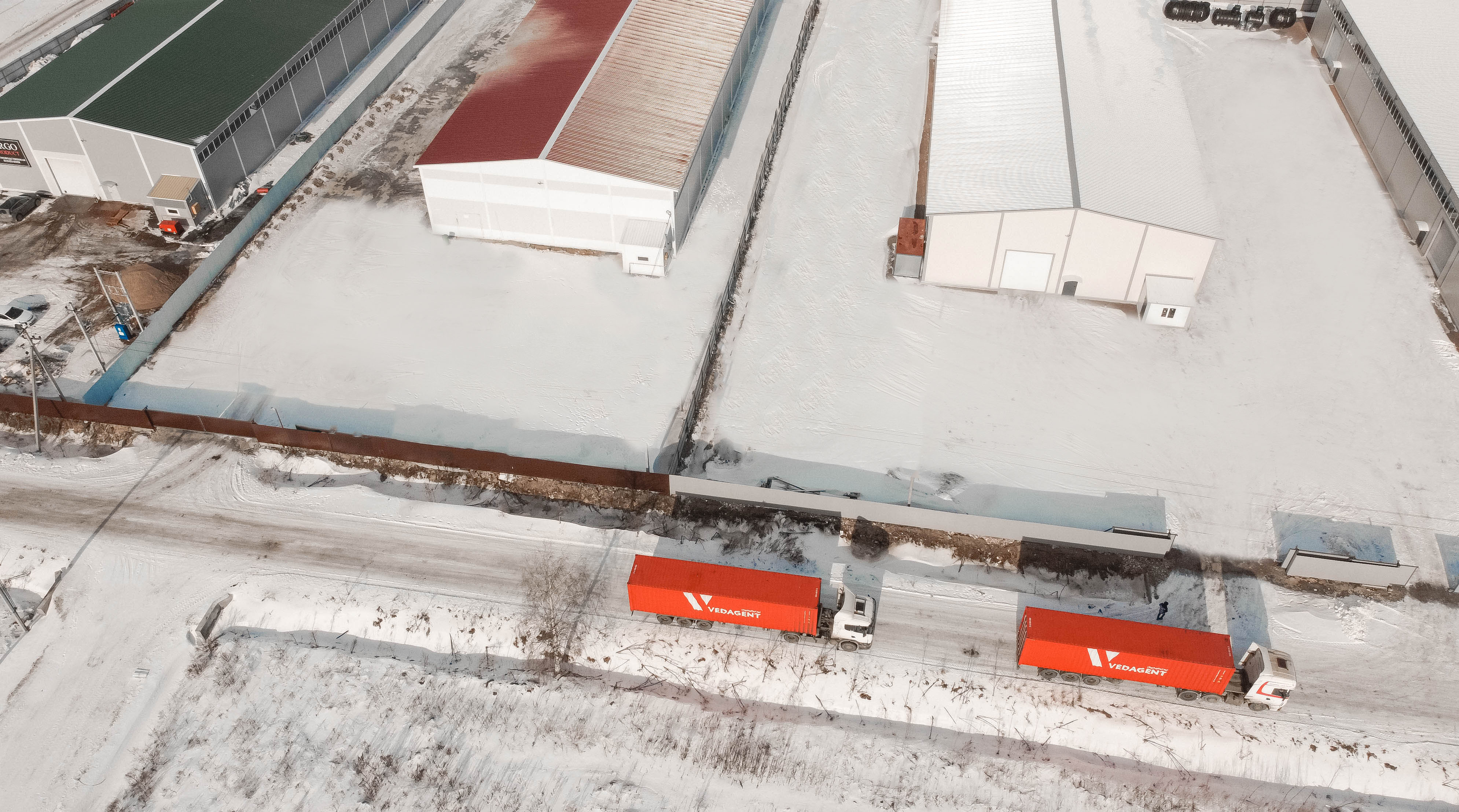 Фотография хорошо убраной дороги в Технопарке Фёдоровское с проезжающими по ней двумя большими ярко оранжевыми фурами. Фото снято зимой с квадрокоптера